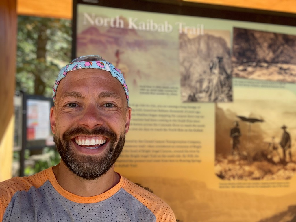 A selfie of Nate Jones smiling broadly with a backwards running hat on. He is seated at the trailhead of the North Kaibab Trail on the North Rim of the Grand Canyon.